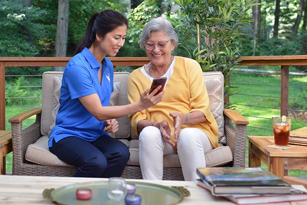 A caregiver in a blue shirt shows an elderly woman something on a phone, both smiling and enjoying a moment together outdoors.