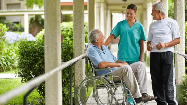A caregiver in teal scrubs chats with two elderly men, one in a wheelchair and the other standing, in a pleasant outdoor setting.