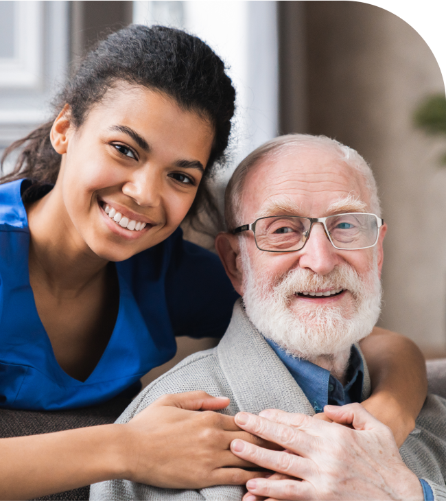 A caregiver and an elderly man smile together, sharing a heartwarming moment on a couch.