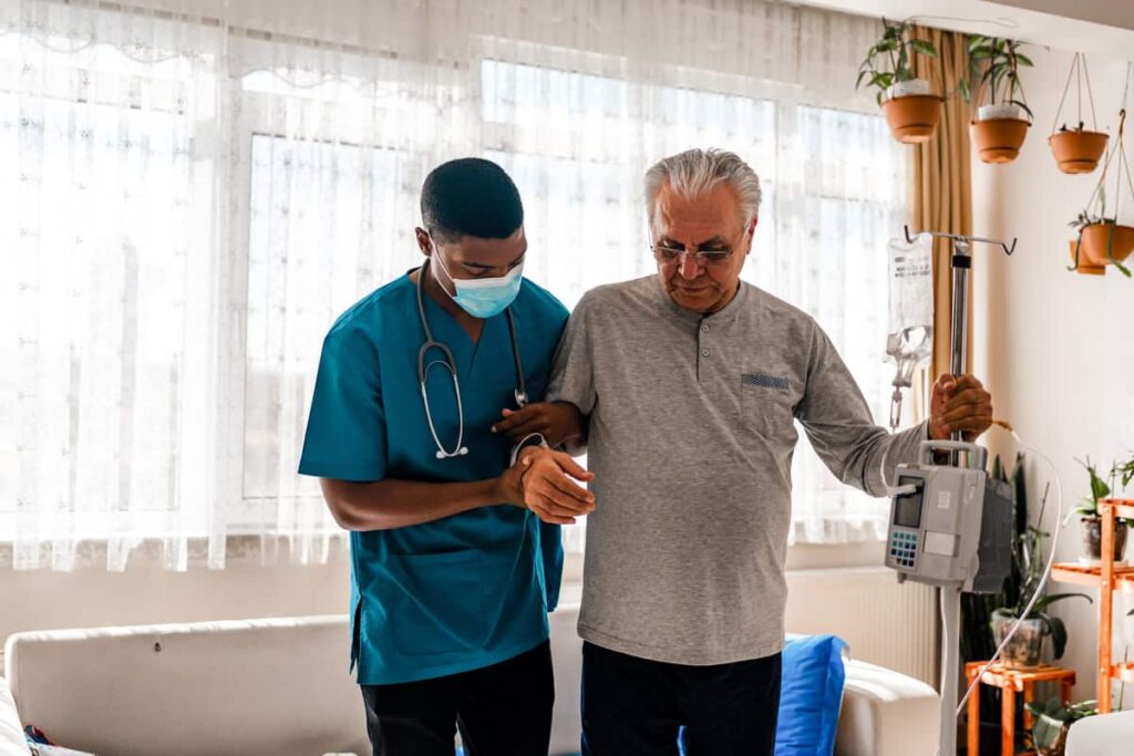 A nurse in blue scrubs assists an elderly man who is standing and holding a portable IV pole in a bright, plant-filled room.