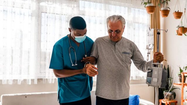 A nurse in blue scrubs assists an elderly man who is standing and holding a portable IV pole in a bright, plant-filled room.
