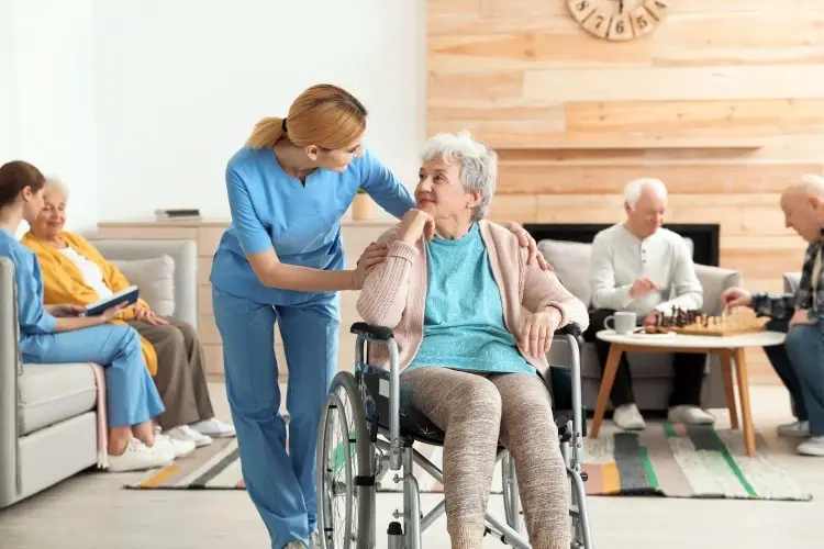 A caregiver in blue scrubs gently assists an elderly woman in a wheelchair while other seniors relax in the background.
