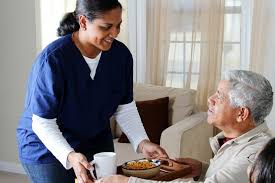A caregiver in blue scrubs serves breakfast and a drink to an elderly man sitting comfortably in a living room.