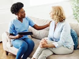 A nurse in blue scrubs sits beside an elderly woman on a couch, placing a comforting hand on her shoulder.
