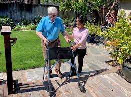 A caregiver supports an elderly man using a walker as they walk outside on a sunny day.