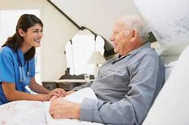 A caregiver in blue scrubs smiles at an elderly man in bed, both sharing a joyful and comforting moment.