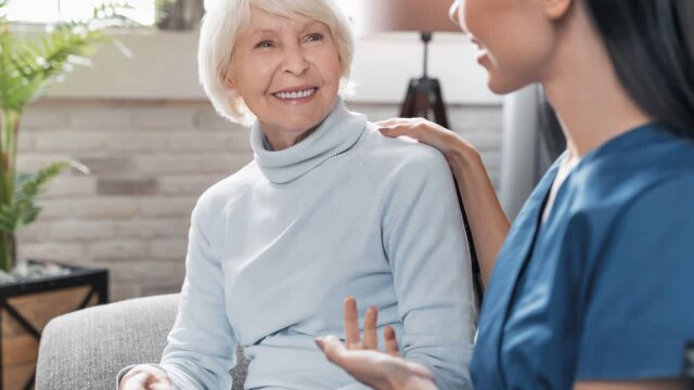 A caregiver offers support to an elderly woman in a cozy living room, helping her with her daily needs.