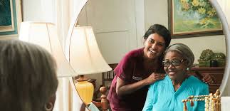 A caregiver and an elderly woman share a joyful moment, as the woman smiles at her reflection in the mirror.