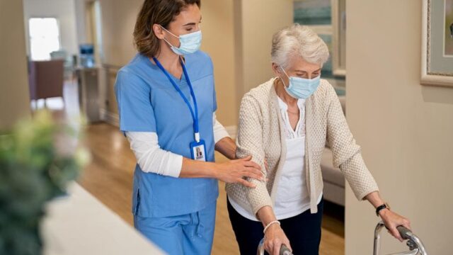 A caregiver in blue scrubs assists an elderly woman with a walker, both wearing masks while walking in a hallway.