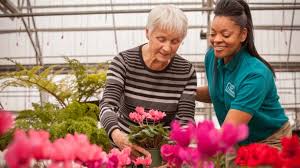 A caregiver in a green shirt helps an elderly woman tend to flowers in a greenhouse, both smiling together.
